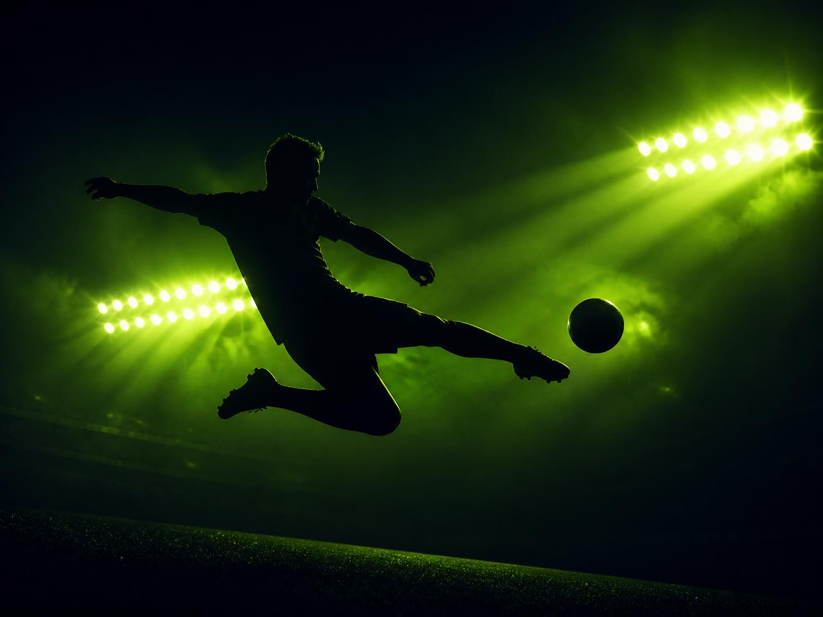 Footballer silhouette striking a ball under stadium floodlights at night